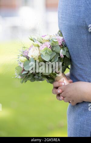 Nahaufnahme der Hand einer Braut mit einem Bouquet aus weißen und rosa Rosen, die die blaue Weste des Bräutigams auf einem Wiesenhintergrund umarmen. Hochwertige Fotos Stockfoto