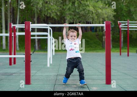 Ein kleiner Junge lernt an einer horizontalen Stange im Freien hochzuziehen. Das Kind hängt an den unebenen Stäben. Stockfoto