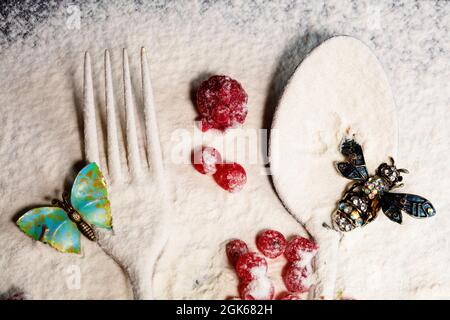 Ein Löffel und eine Gabel, rote Beeren sind mit Mehl bestreut und eine Broschüre in Form von Insekten liegt oben. Kreative Zubereitung von Speisen. Stockfoto