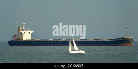 Cascais, Portugal; 31. August 2021: Kleines Segelschiff, das neben einem riesigen Massengutfrachter vor der Küste von Cascais (Portugal) vor Anker liegt Stockfoto