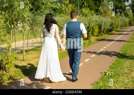 Das Brautpaar läuft den Parkweg entlang und hält sich die Hände. Ein Mann und ein Mädchen machen einen romantischen Spaziergang. Im Sommer spazieren Liebhaber im Park. Stockfoto