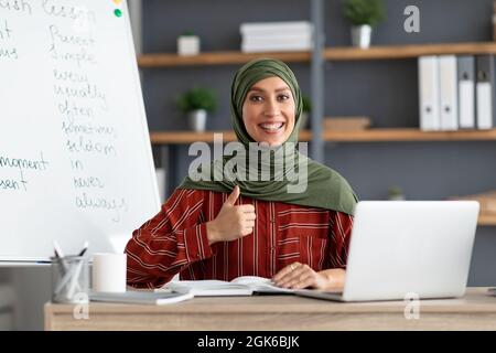 Islamischer Lehrer im Kopftuch, der am Schreibtisch sitzt und den Daumen nach oben zeigt Stockfoto