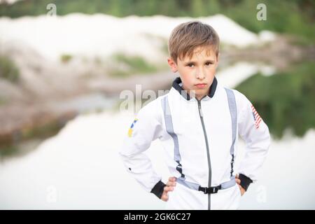 Ein hübscher Junge im weißen Anzug eines amerikanischen Astronauten blickt vor dem Hintergrund der Natur in die Kamera. Stockfoto