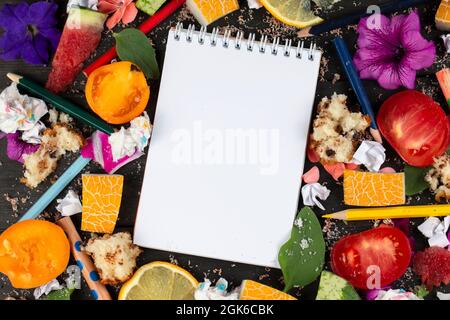 Papiernotizbuch mit leeren Seiten, zerknitterten Papierkugeln und Buntstiften, verstreuten Lebensmitteln auf Holzhintergrund, kreatives Kochrezept-Schreibkonzept. Kommi Stockfoto