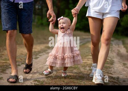 Ein glückliches, einjähriges Mädchen wird von Vater und Mutter festgehalten. Kleines Kind, das mit den Eltern läuft. Ein süßes Baby lernt mit Hilfe ihrer Eltern zu gehen. Stockfoto