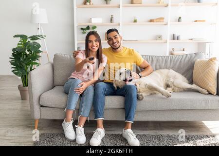 Junges liebevolles multirassisches Paar, das mit Hund auf der Couch sitzt und zu Hause Fernsehen schaut Stockfoto