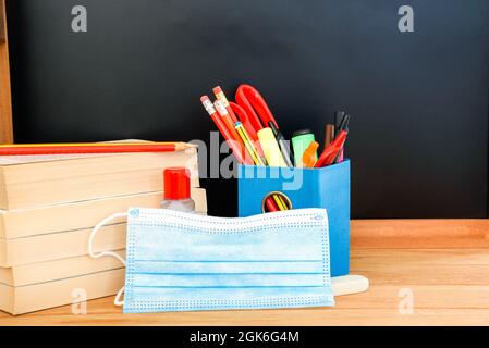Zurück in die Schule 2021: Lehrbücher, Stifte, Bleistifte, eine Gesichtsmaske und eine Flasche Desinfektionsmittel auf einem Holzschreibtisch mit einer leeren Tafel im Hintergrund Stockfoto
