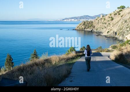 Frau mit blonden Haaren geht eine kleine Straße hinunter zum Strand von Cañuelo von Nerja mit einem Wachturm und der Stadt Nerja im Hintergrund. Maro Cerro Stockfoto