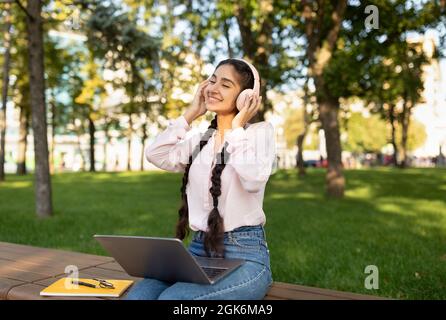 Fröhliches indisches Studentenmädchen, das sich auf dem Campus mit einem Laptop ausruhte, Kopfhörer trug und Musik hörte, auf der Bank saß Stockfoto