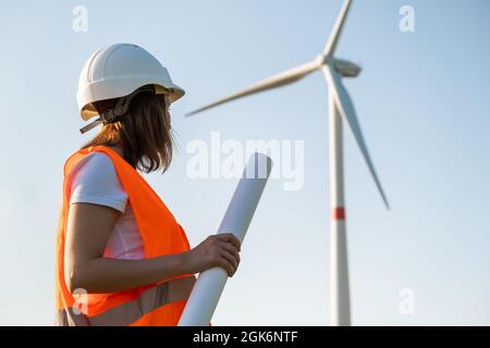Ingenieur in Hut und Schutzweste hält vor dem Hintergrund von Windmühlen ein Planprojekt für den Bau von Windturbinen im Feld Stockfoto