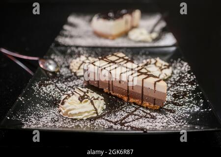 Portion Kuchen mit drei Pralinen auf einem Teller und einem schwarzen Hintergrund, bestreut mit Puderzucker, mit Creme und Schokoladensirup Stockfoto