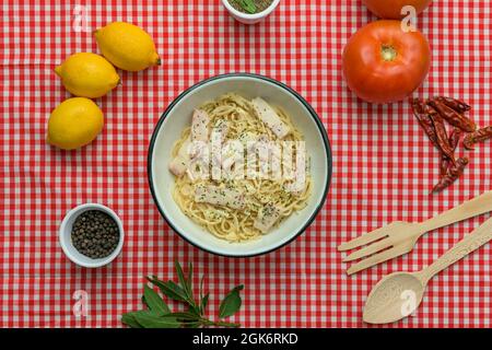 Schüssel mit Spaghetti Carbonara mit Speck auf dem Tisch mit rot-weißer Tischdecke. Holzbesteck, Tomaten, Cayenne, Zitronen, Pfeffer und Lorbeerblatt Stockfoto