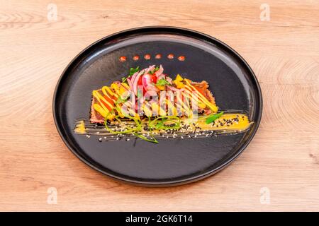 Schwarzer Teller mit rotem Thunfisch-Tataki mit Saucen, Samen, Zwiebeln und Granatapfelkörnern auf Eichenholzhintergrund Stockfoto