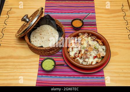 Mexikanisches Rindfleisch alambre Rezept mit viel geschmolzenem Käse, Guacamole und Maulwurfsauce, Mais-Tortillas in Ton und Weidenbehälter Stockfoto