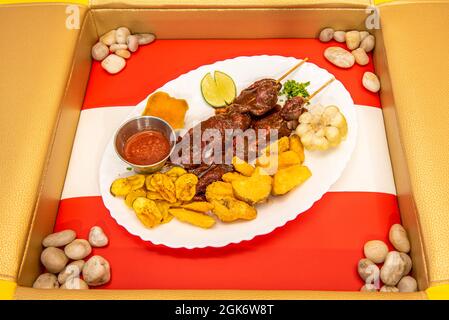 Weißes Tablett auf der Flagge Perus mit Kochbananen in Chips, Bratkartoffeln und Kartoffeln, die zwei Pinchos peruanischer Anticuchos auf dem Grill begleiten Stockfoto