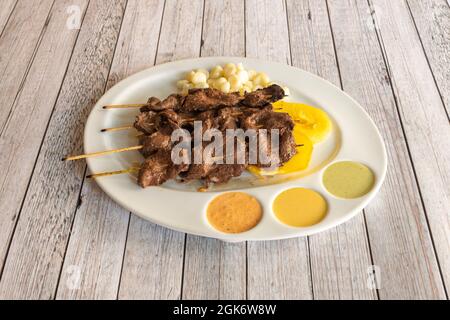 Tablett von peruanischen Anticuchos auf Holzstäbchen auf dem Grill mit gekochtem Mais, Kartoffeln und Süßkartoffeln mit Saucen zum Eintauchen gekocht punktiert Stockfoto