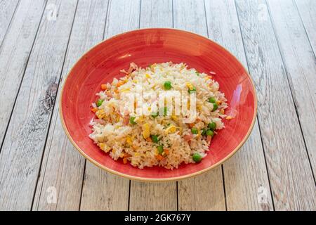Drei köstliche Reisgerichte, typisch für ein chinesisches Restaurant, mit gebratenen Eierfilamenten, Karottenteilen, Schinken und Erbsen auf einem roten, tiefen Teller auf einem weißen Tisch Stockfoto