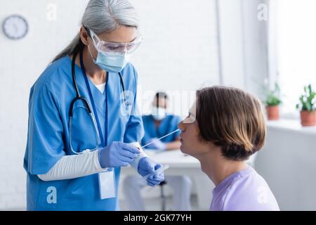 Reifer asiatischer Arzt in medizinischer Maske und Brille, die Coronavirus-Test für jungen Mann macht Stockfoto