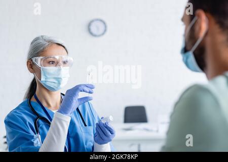 Reifer asiatischer Arzt in medizinischer Maske und Brille Vorbereitung Spritze mit Impfstoff in der Nähe verschwommener Mann Stockfoto