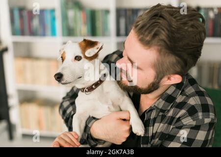 Glücklicher männlicher Besitzer von Jack russell Terrier Hund, fühlt sich verantwortlich für die Pflege von Haustier, vor Bücherregalen Hintergrund stehen. Menschen und Stockfoto