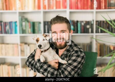 Glücklicher männlicher Besitzer von Jack russell Terrier Hund, fühlt sich verantwortlich für die Pflege von Haustier, vor Bücherregalen Hintergrund stehen. Menschen und Stockfoto