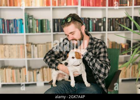 Glücklicher männlicher Besitzer von Jack russell Terrier Hund, fühlt sich verantwortlich für die Pflege von Haustier, vor Bücherregalen Hintergrund stehen. Menschen und Stockfoto