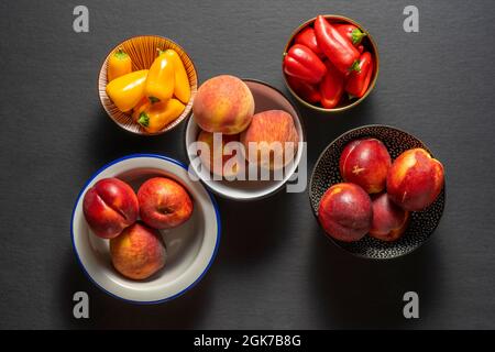 Verschiedene Schalen mit Nektarinen, Pfirsichen und gelben und anderen roten Paprika auf schwarzem Hintergrund Stockfoto