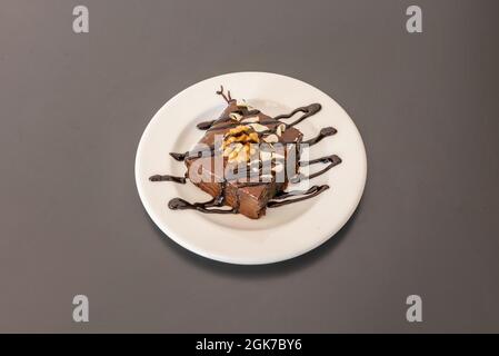 Bunte Dessertportion Schokoladenbrownie-Kuchen mit getrockneten Früchten und Nüssen, Linien von Schokoladensirup und weißen Schokoladenspangen auf einem weißen Teller Stockfoto