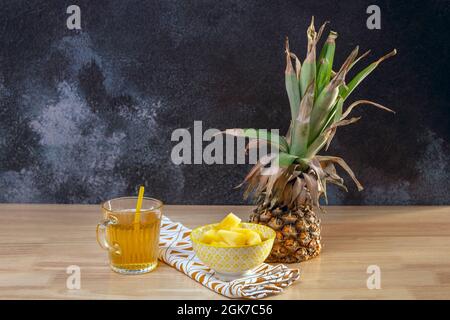 Eine halbe Ananas auf dem Tisch, eine gelbe Schüssel voller Ananasbrocken und ein Glas mit Stroh und Ananassaft Stockfoto