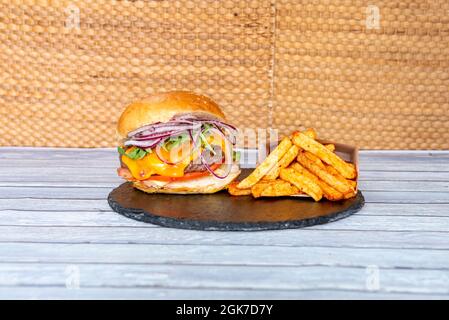 Köstlicher Rindfleisch-Burger mit geschmolzenem Cheddar-Käse, rohen roten Zwiebelringen, Arugula-Blättern, Tomatenscheiben, Senfsauce und Paprika-Fries Stockfoto