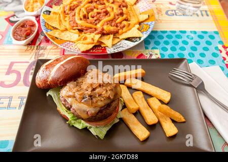 Rindfleisch-Burger mit vielen geschmorten Zwiebeln, Toast, Eisbergsalat, geschmolzenem Käse und Tomaten, serviert mit pommes frites an einem Restauranttisch mit Chili con Stockfoto