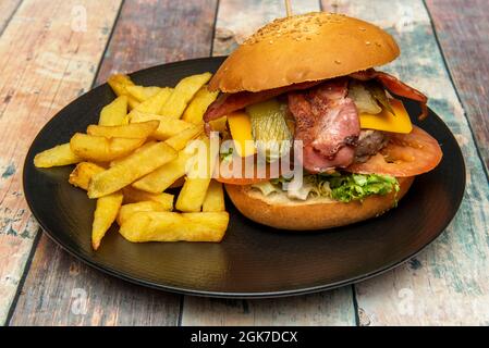 Angus-Rindfleisch-Burger mit Gurken, Cheddar-Käse, gebratenem Speck, Tomatenscheiben, Salat und Pommes frites Stockfoto