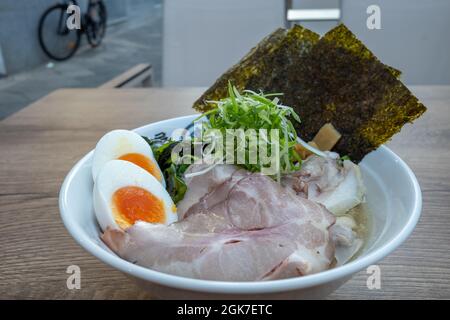 Shio Ramen, japanische Nudellattenmuscheln-Suppe, darauf Schweinefleisch, gekochtes Ei, Bambussprossen, rote Zwiebel und Nori, auf einem hölzernen Außentisch. Stockfoto