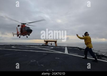 Ens. Ryan Dunkle, ein Crewmitglied der Küstenwache, Cutter Healy (WAGB 20), übermittelt Landesignale an Piloten der Air Station Kodiak, die während des gemeinsamen Flugtrainings vor der Küste Alaskas während des Einsatzes von Healy in der Arktis am 25. August 2021 einen MH-65 Dolphin fliegen. Die Landeanzeiger sind für die Kommunikation mit der Flugbesatzung und den Cutter-Besatzungsmitgliedern während des Flugbetriebs verantwortlich. Stockfoto