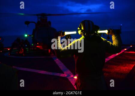 Ens. Ryan Dunkle, ein Crewmitglied der Küstenwache, Healy (WAGB 20), gibt während des nächtlichen gemeinsamen Flugtrainings mit einer Flugmannschaft der Air Station Kodiak Kommandos an ein Verzurrungsteam, während Healys Einsatz in der Arktis am 25. August 2021 vor der Küste Alaskas. Die Landeanzeiger sind für die Kommunikation mit der Flugbesatzung und den Cutter-Besatzungsmitgliedern während des Flugbetriebs verantwortlich. Stockfoto