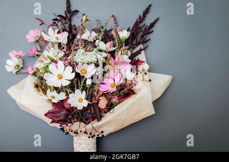 Frisches Bouquet von weißen rosa Blüten mit burgunderroten Blättern, eingewickelt in Papier und auf grauem Hintergrund angeordnet. Kosmos kombiniert mit wilden schwarzen Beeren Stockfoto