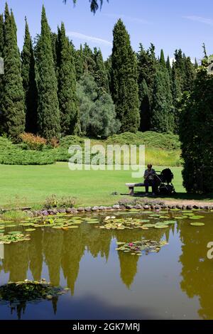 Großmutter mit Kinderwagen, die an einem sonnigen Tag in der Nähe eines kleinen Teiches sitzt. Sigurtà Garden Park, Valeggio sul Mincio, Venetien, Italien. Vertikale Aufnahme. Stockfoto