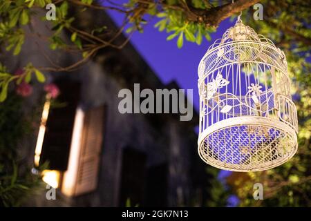 Dekorativer Vogelkäfig, der nachts im Garten an einem Baum hängt. Schäbiges, ländlich-schickes Wohndekor, Idee zur Garteninspiration. Stockfoto