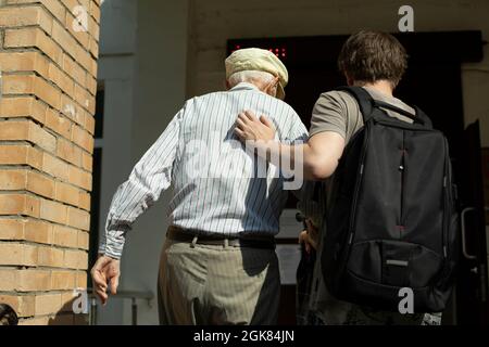 Der Enkel hilft Opa zu Fuß zum Eingang des Krankenhauses. Ein Freiwilliger führt einen Rentner an, um sich impfen zu lassen. Ein Sozialarbeiter begleitet einen alten ma Stockfoto