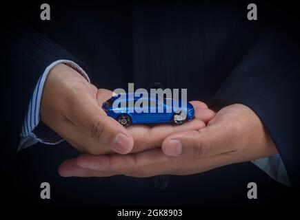 Asiatischer Geschäftsmann zeigt ein Spielzeugauto auf seiner Handfläche. Konzeptioneller Ideeneigentum, Versicherung und Transport. Stockfoto