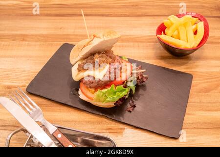 Kalbsburger auf den Punkt mit Tomaten und Salat mit Ketchup und Senf, geschmolzenem Manchego-Käse und karamellisierten Zwiebeln mit Bratkartoffeln, Messer und Stockfoto