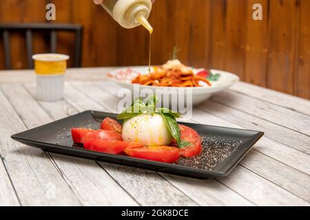 Olivenöl mit einer Flasche auf einen Burrata-Käseball mit Tomatenscheiben auf einen weißen Tisch gießen Stockfoto