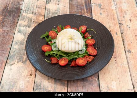 Kugel aus italienischem Burrata-Käse mit Tropfen Olivenöl und auf einem Bett aus Rucola und Krone aus Kirschtomaten und getrockneten Tomaten Stockfoto