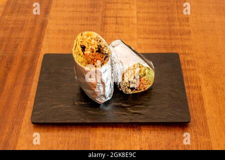 Zerfetzter Rinderburrito mit Salat und schwarzen Bohnen, weißem Reis und Weizentortilla in Aluminiumfolie auf einem schwarzen Schiefertablett verpackt Stockfoto