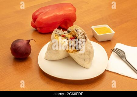 Köstlich aussehender mexikanischer Burrito mit schwarzen Bohnen, weißem Reis, Fleisch und Saucen auf dem Tisch mit Salsa, rotem Pfeffer und roter Zwiebel Stockfoto