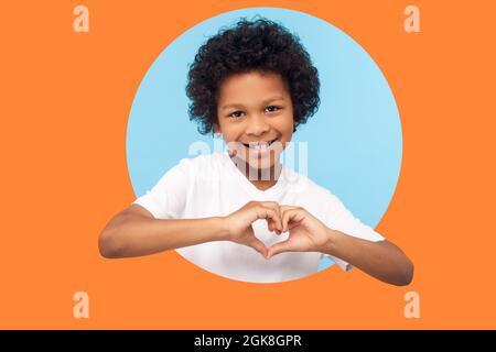 Porträt eines lächelnden kleinen Jungen mit lockigen Haaren in weißem T-Shirt, das mit den Fingern in Herzform zeigt und unschuldige Kindheitsliebe zum Ausdruck bringt Stockfoto