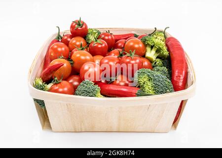 Feiner, ungelackter Holzkorb, gefüllt mit Kirschtomaten, grünen Brokkoli-Stücken und roten, heißen Chilischoten auf weißem Hintergrund Stockfoto