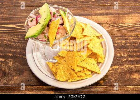 Peruanischer Fisch-Ceviche mit einer Scheibe Avocado in einem Glas montiert und mit Nachos auf einem weißen Teller garniert Stockfoto