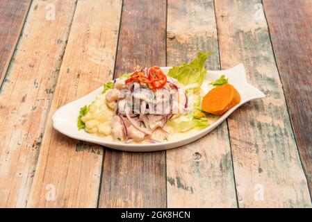 Peruanische corvina ceviche, mariniert mit Limette, Zwiebeln, Maiskörnern, Süßkartoffeln und Salat auf einem fischförmigen Teller auf einem Holztisch Stockfoto