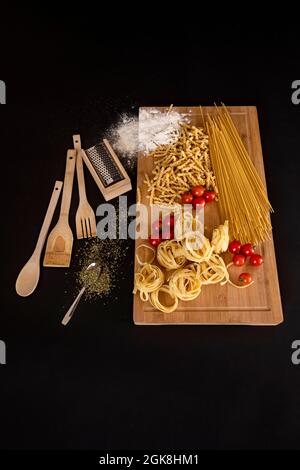 Küchengeräte aus Holz, frische rohe italienische Pasta, Kirschtomaten, Orangano, Weizenmehl und Bambustisch und schwarzer Hintergrund Stockfoto